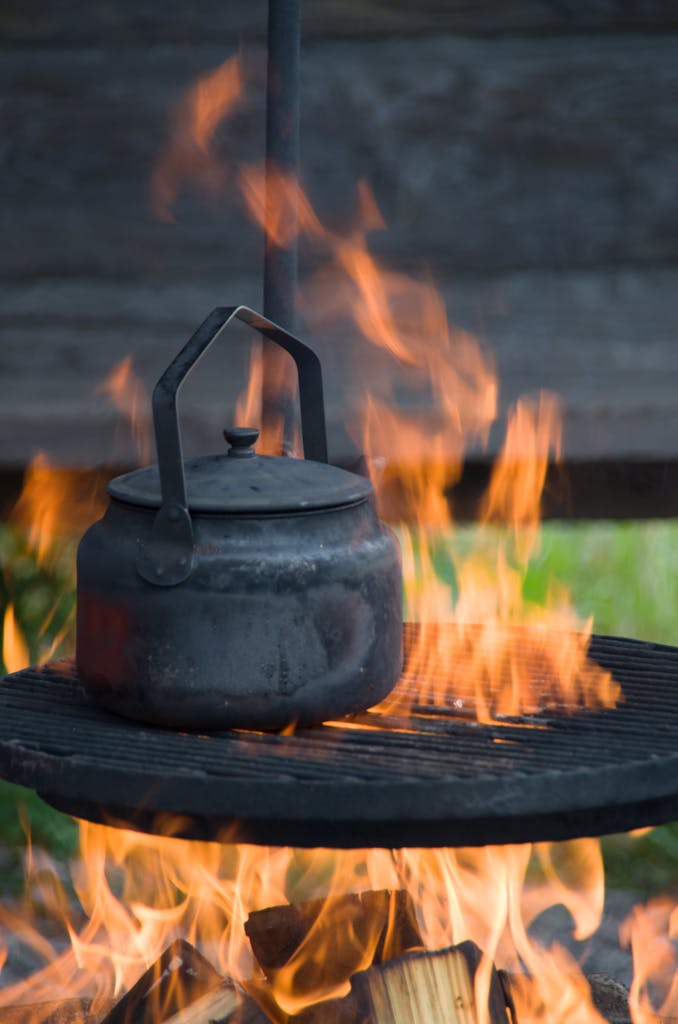 A kettle boiling over an outdoor campfire, surrounded by flames, creating a rustic cooking scene.